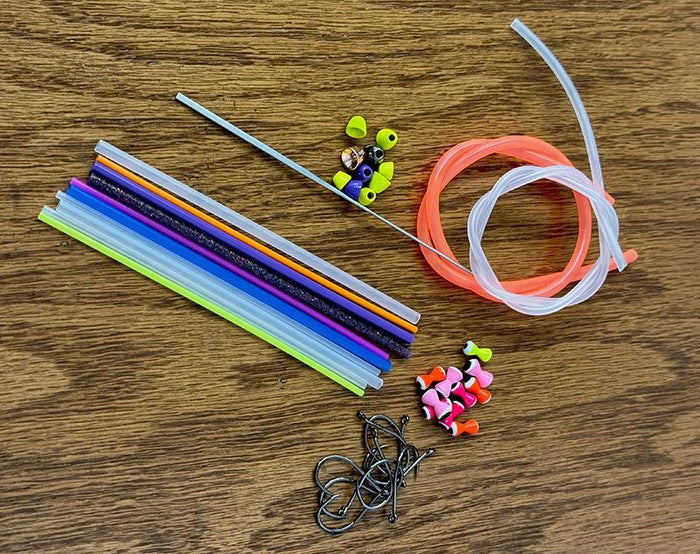 Aqua Flies Tube Kit displayed on a wood surface featuring colorful plastic tubes, assorted fishing hooks, silicone tails, and metal needles for assembling custom fishing lures.