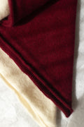 A close-up of two folded cashmere triangle scarves, showing a deep burgundy red outer layer and a cream inner layer on a light textured background.