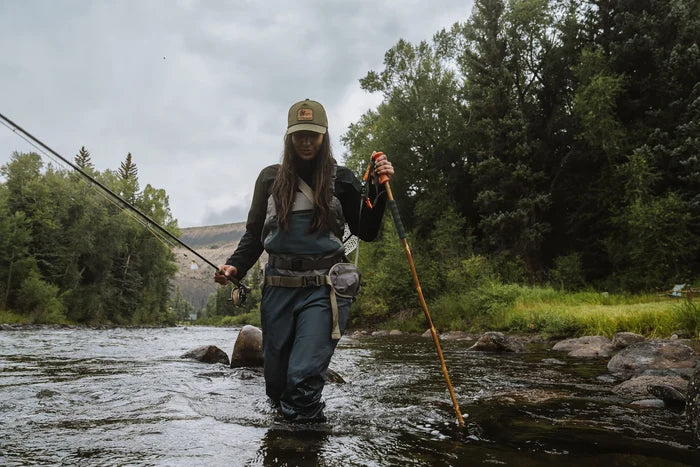 Woman wearing fishing waders and a cap standing in a river holding a fishing rod in one hand and a Fishpond Grass Stick Wading Staff in the other for stability while fishing.