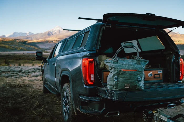 Fishpond Horse Thief Tote in green canvas with multiple compartments and durable handles, sitting on the open tailgate of a black pickup truck in an outdoor landscape.