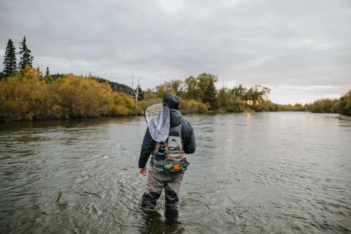 Angler standing in a river wearing waders and carrying a net, shown from behind wearing the Fishpond Stormshadow Lumbar Pack with fishing gear attached.