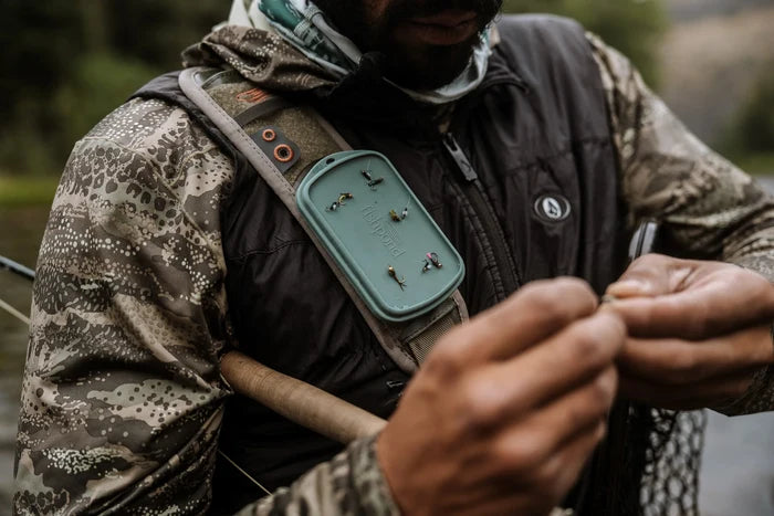 Angled view of a person wearing a camo jacket and a dark vest with a Fishpond Tacky Grande Fly Dock MagPad attached to their chest strap holding several fishing flies, preparing fishing gear outdoors near a river.