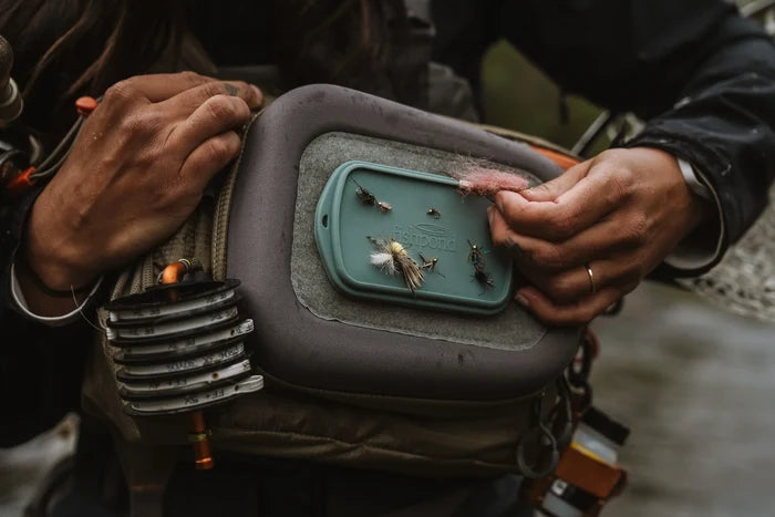 Close-up of a person holding a fishing vest equipped with the Fishpond Tacky Grande Fly Dock MagPad displaying several fishing flies attached, showcasing its soft magnetic surface designed for convenient fly fishing tackle storage and quick access.