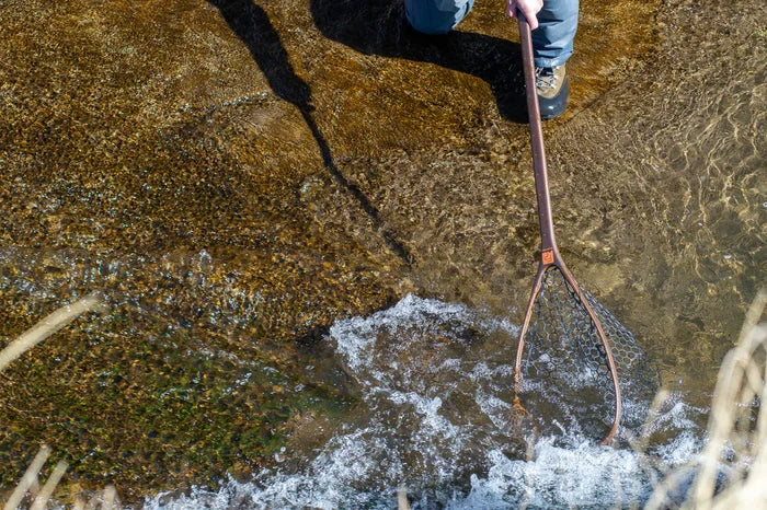 Person standing in shallow water holding a Fishpond Yampa Guide Net 2.0 with a wooden frame and rubber mesh designed for catching fish safely without harming them.