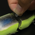 Close-up of a hand applying Galápagos Swim Bait Joint Pads to the joint of a segmented fishing lure, highlighting detailed texture and vibrant green and blue colors.