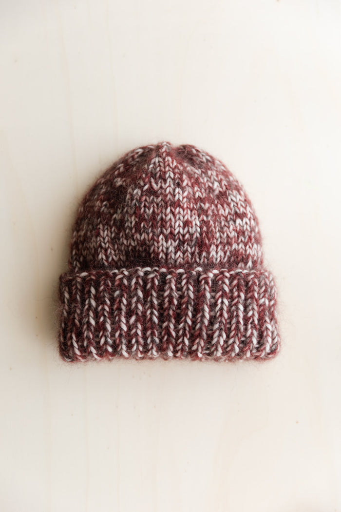 A hand-knit beanie hat in a marled mix of burgundy, brown, and cream wool, photographed on a warm off-white surface.