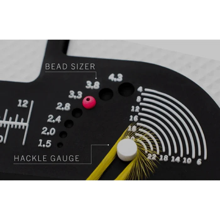 Close-up of a black MFC Fly Tying Mat section featuring bead sizer holes with measurements, a pink bead in one hole, hackle gauge markings, and a feather secured by a white pin.