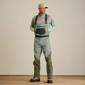 Man wearing Orvis Clearwater Men's Wader in muted green tones with adjustable black suspenders, front pocket, and black belt, paired with hiking boots and a beige cap.