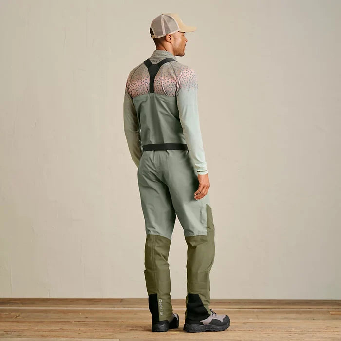 Man wearing Orvis Clearwater Men's Wader in muted green with darker reinforced knee and ankle panels, paired with a light long-sleeve shirt and beige cap, shown from the back standing on wooden floor.