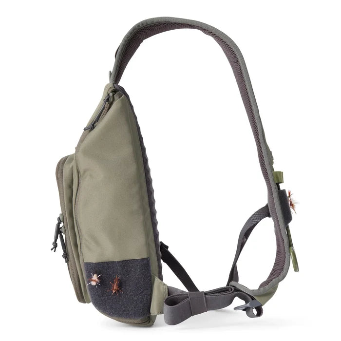 Orvis Mini Sling Pack in olive green with adjustable padded strap, featuring multiple zippered compartments and fly fishing flies attached to the front panel.