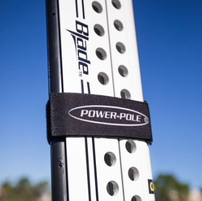 Close-up of a black Power-Pole Travel Strap For All Power-Pole Models SKU:TRAVEL-STRAP-F securing a white fishing tackle blade against a clear blue sky background.