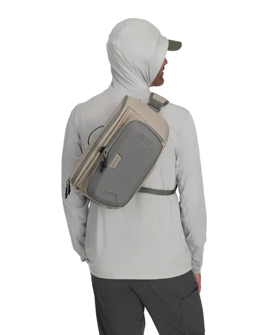 Man wearing light gray hooded jacket and dark pants carrying a beige and gray Simms Headwaters Sling Pack across his back with adjustable straps and multiple zippered compartments.