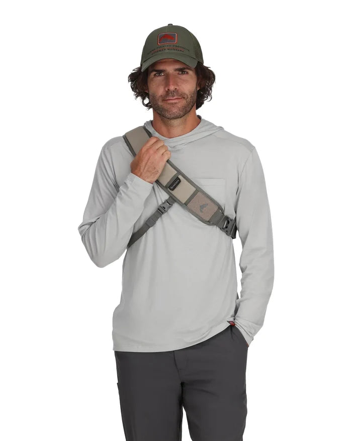 Man wearing a gray long-sleeve shirt and olive green cap, modeling a tan and gray Simms Headwaters Sling Pack worn diagonally across his chest, designed for fishing tackle storage.
