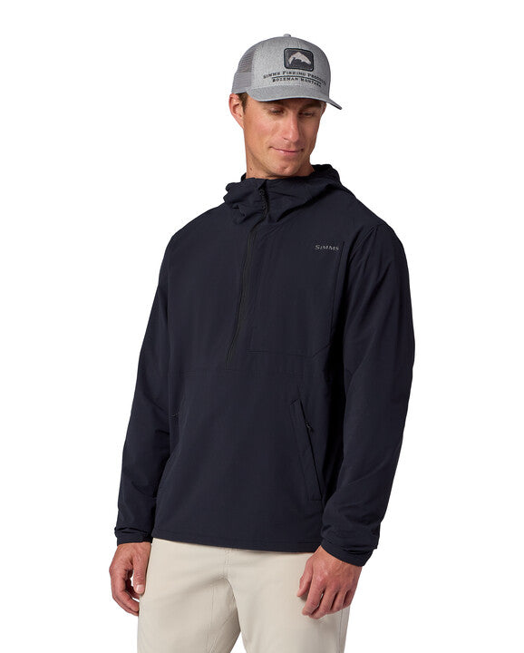 A man wearing a black Simms Rogue Flex Half-Zip Pullover with a hood and front zip, paired with beige pants and a gray baseball cap, standing and looking down.