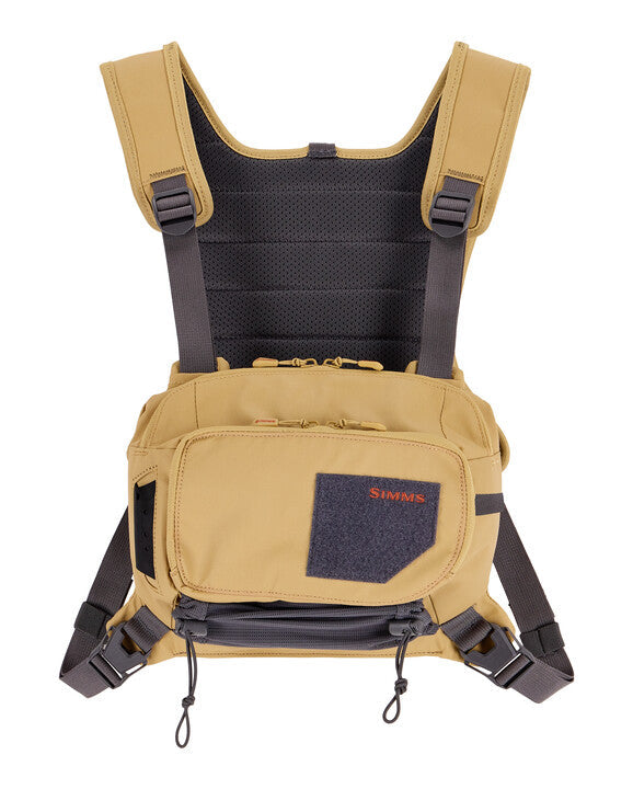 Simms Tributary Chest Pack in tan with padded shoulder straps, multiple zippered compartments, adjustable buckles, and breathable mesh back panel designed for fishing tackle storage and comfort.