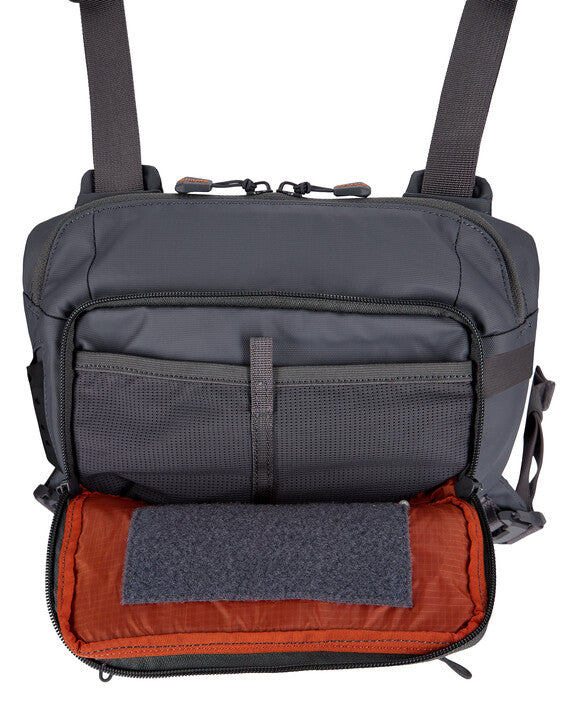 Simms Tributary Chest Pack in dark gray with adjustable straps, multiple zippered compartments, and an orange-lined interior designed for organizing fishing tackle and accessories.