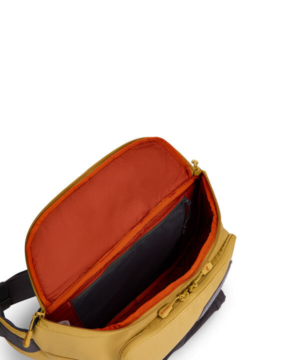 Simms Tributary Hip Pack shown open highlighting spacious interior with multiple compartments, durable beige exterior, and contrasting orange lining designed for organized fishing tackle storage.