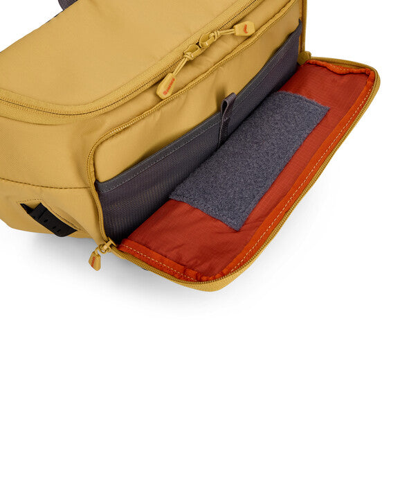Simms Tributary Hip Pack in tan, shown open to reveal multiple compartments including a zippered pocket, mesh organizer, and an orange interior lining.
