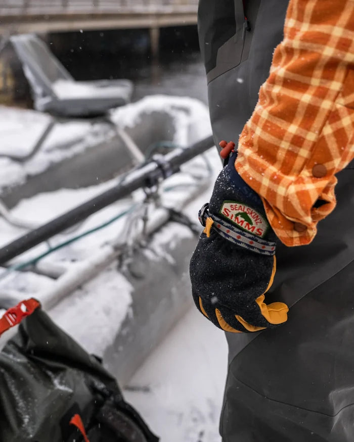 Close-up of a person wearing an insulated Simms X Sealskinz Baxter Glove with waterproof and breathable fabric, standing outdoors near fishing gear in a snowy environment.