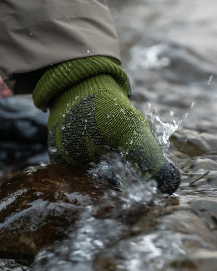 Close-up of a hand wearing a green Simms X Sealskinz Rangeley Waterproof Glove, partially submerged in flowing water, demonstrating its water-resistant and textured grip features.