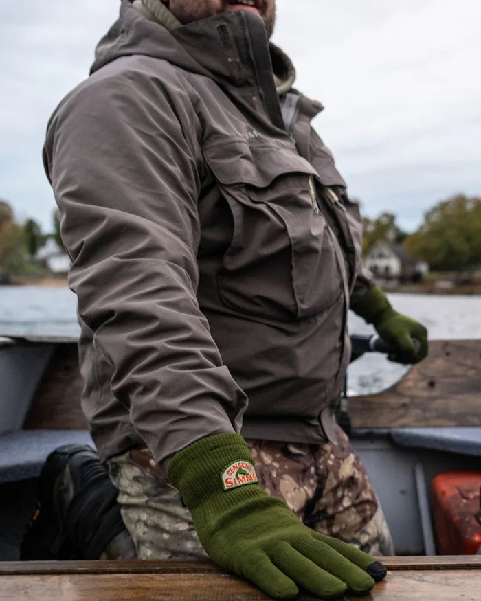 Man wearing olive green Simms X Sealskinz Rangeley Waterproof Glove while sitting on a boat, dressed in a brown jacket and camo pants near a lake.