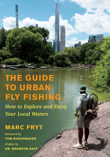 Cover of "The Guide To Urban Fly Fishing" showing an angler standing on a rocky shore casting a fly fishing rod into a calm urban waterway with city skyscrapers and green trees in the background.