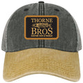 Thorne Bros Dashboard Legacy Trucker Hat featuring a two-tone design with a grey fabric crown, beige distressed bill, and a front patch logo displaying a musky fish and custom rod and tackle branding.