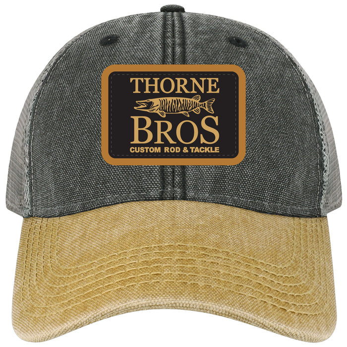 Thorne Bros Dashboard Legacy Trucker Hat featuring a two-tone design with a grey fabric crown, beige distressed bill, and a front patch logo displaying a musky fish and custom rod and tackle branding.