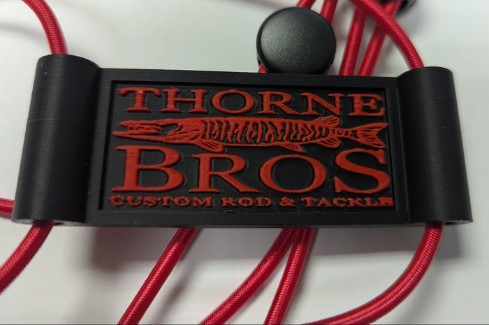 Close-up of a Thorne Bros Graph Cover Strap in black with red lettering and logo, featuring durable red cords, designed for securing fishing rods and tackle.