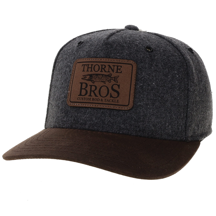 Thorne Bros. Wool Cut Above Hat featuring a dark gray wool crown and a brown suede curved brim with a leather patch logo representing a musky fish and custom rod and tackle branding.