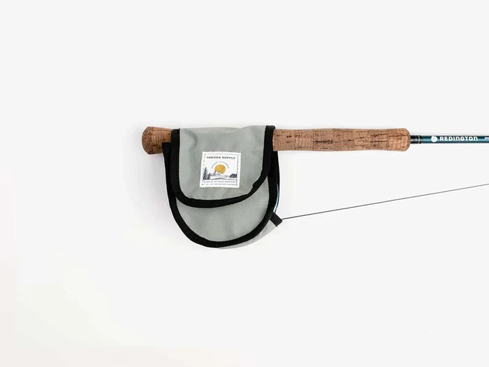 Yakoda Reel Cover in silver gray fabric with black trim, designed to protect fishing reels, shown attached to a fly fishing rod with cork handle.